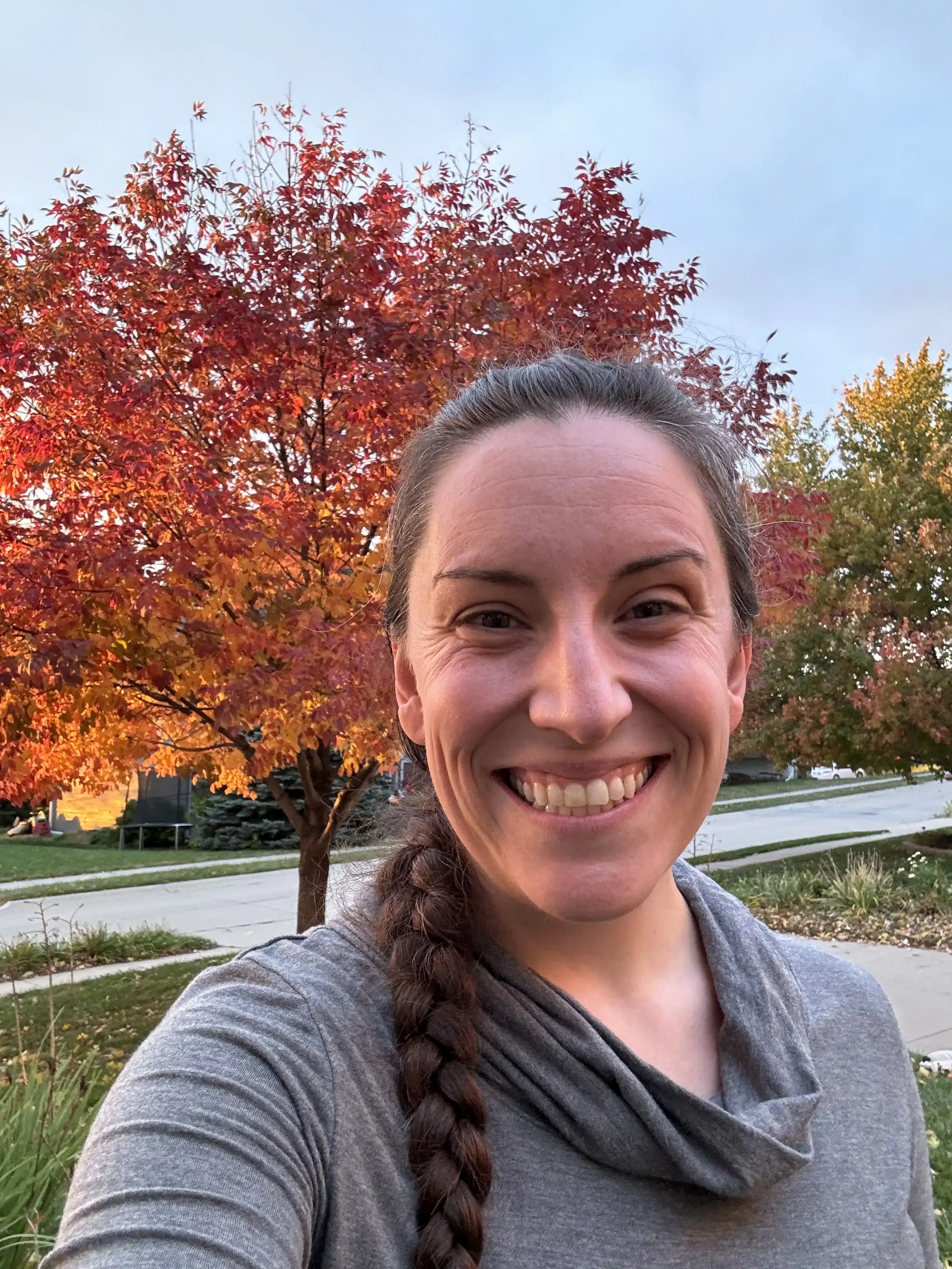 Diane Newcomer - Read-Aloud Revival ® with Sarah Mackenzie Smiling woman taking a selfie outdoors with vibrant red and orange autumn trees behind her.