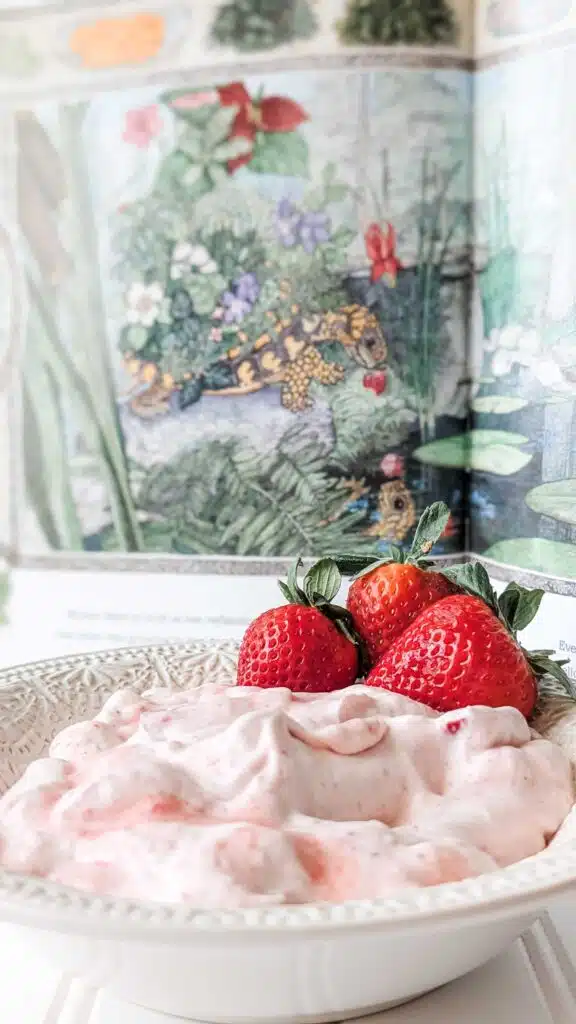 Mossy Strawberry - Read-Aloud Revival ® with Sarah Mackenzie Close-up of a pink strawberry dessert with fresh strawberries on top, served in a white bowl on a floral background.