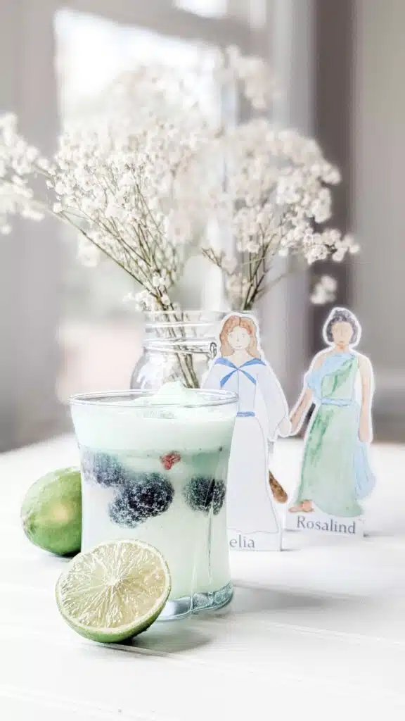 Woodland Spritzer 1 - Read-Aloud Revival ® with Sarah Mackenzie Mint-green drink in a glass with blueberries and lime, on a white surface with delicate white flowers in a jar and paper cutout figures in the background.