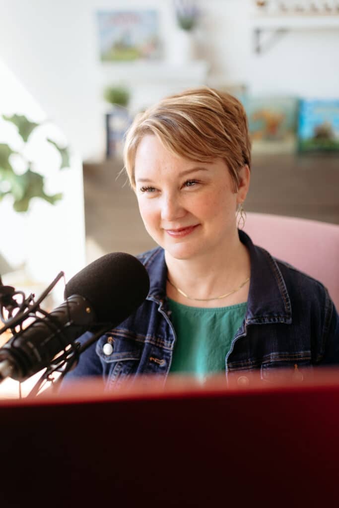 Smiling woman in a denim jacket speaking into a large microphone in a recording studio.