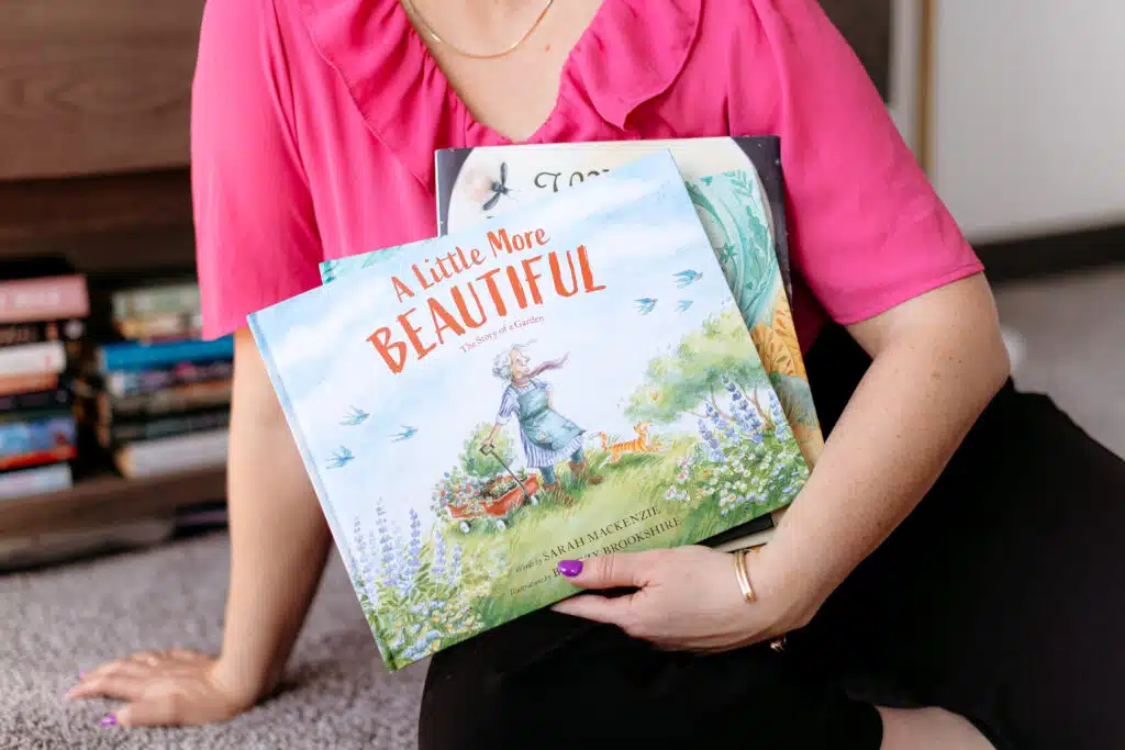 sarah_mackenzie-33 - Read-Aloud Revival ® with Sarah Mackenzie Person in a pink blouse sits on a carpet, hugging a colorful children’s book titled 'A Little More Beautiful' with illustrations visible on the cover.