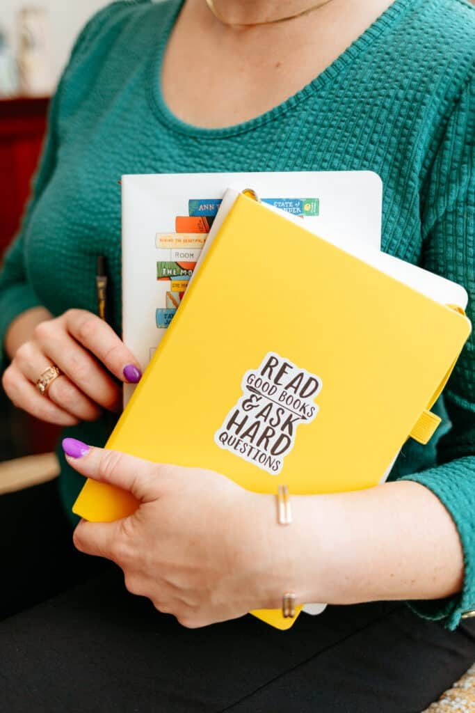 Person in a teal sweater holds a bright yellow notebook over a colorful stack of books, with a sticker that says 'Read good books & ask hard questions'