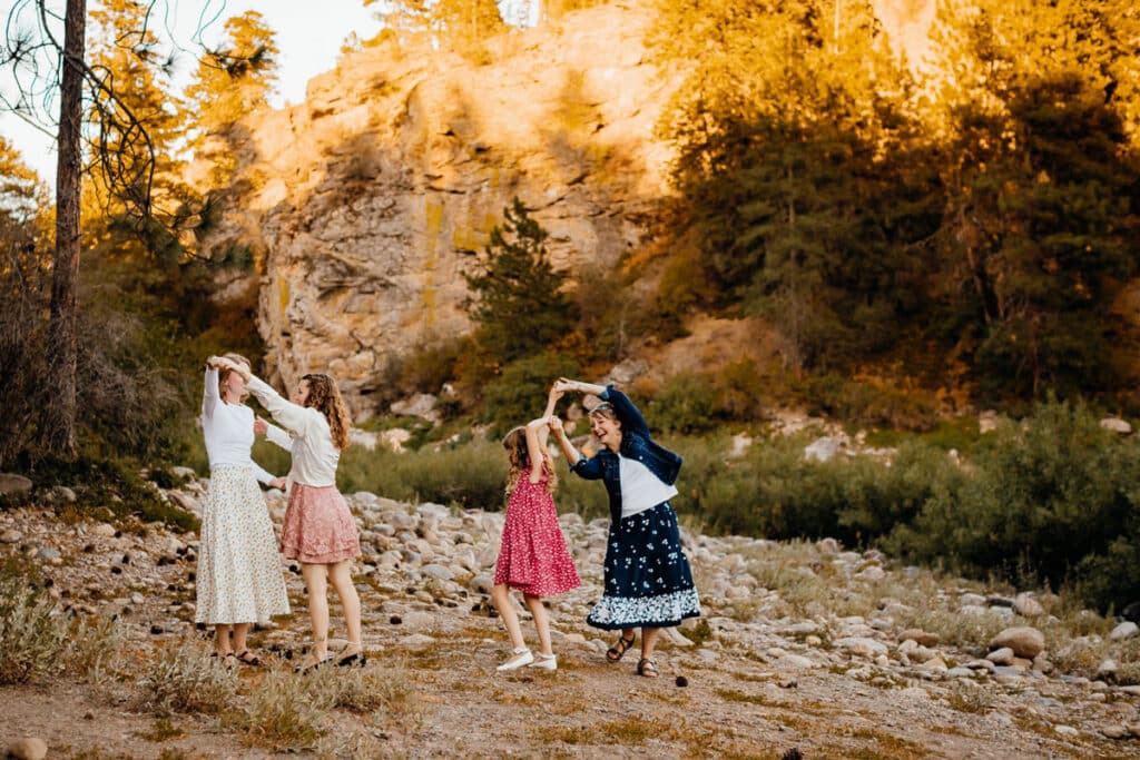 Four girls dancing hand-in-hand in a rocky canyon at sunset, wearing long floral skirts and dresses while the warm light hits the cliff.