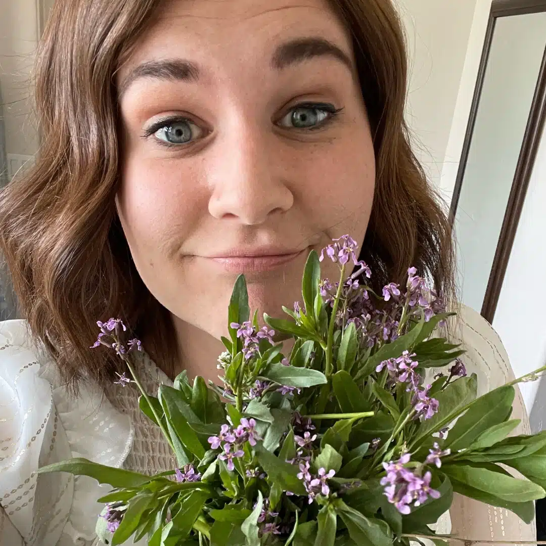 Woman smiling with a bouquet of small purple flowers close to her face in a bright room, wearing a white top.