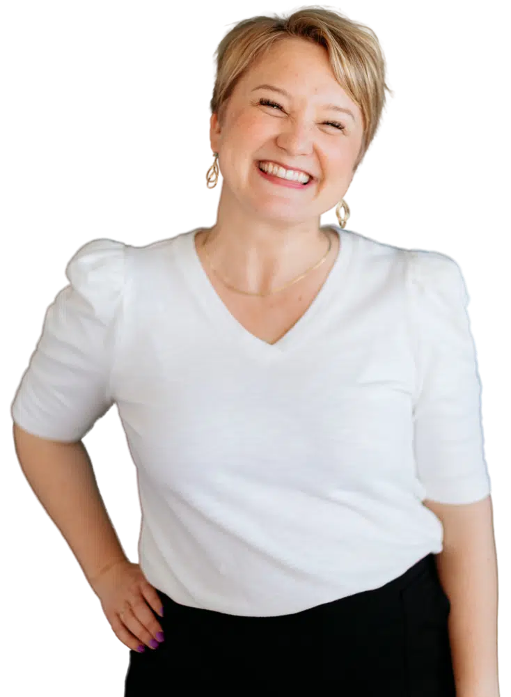 Smiling woman with short blonde hair, wearing a white V-neck shirt and gold earrings, standing with a hand on her hip.