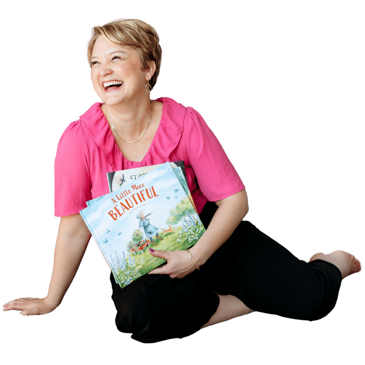 Smiling woman in a pink blouse sits on the floor, holding a children's book titled A Little More Beautiful.