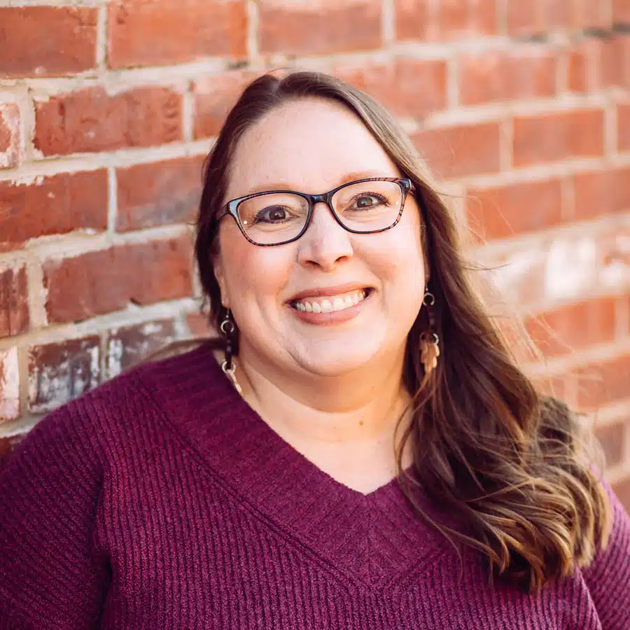 Angie-Dubbert - Read-Aloud Revival ® with Sarah Mackenzie Smiling woman with glasses in a maroon knit sweater poses against a brick wall.