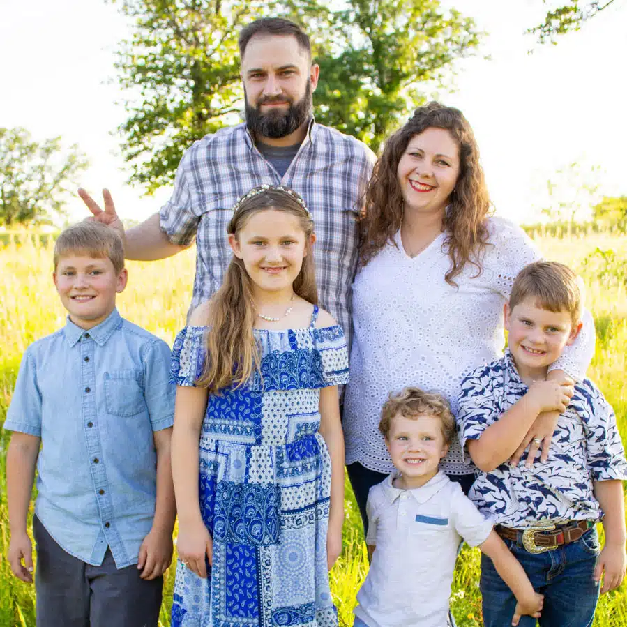 Jamie-Braden - Read-Aloud Revival ® with Sarah Mackenzie Family of six posing in a sunlit field, smiling at the camera: father, mother, a girl in blue dress, and three boys.
