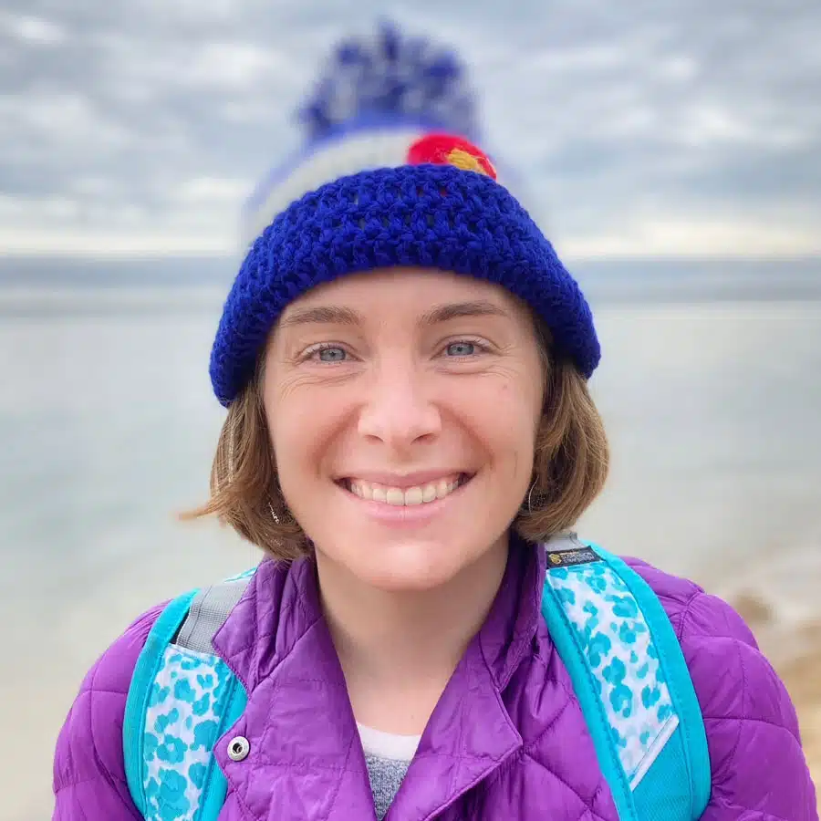 Robyn-Olear - Read-Aloud Revival ® with Sarah Mackenzie Smiling woman in a blue knit beanie and purple jacket at the beach, with a teal backpack visible on her shoulders.