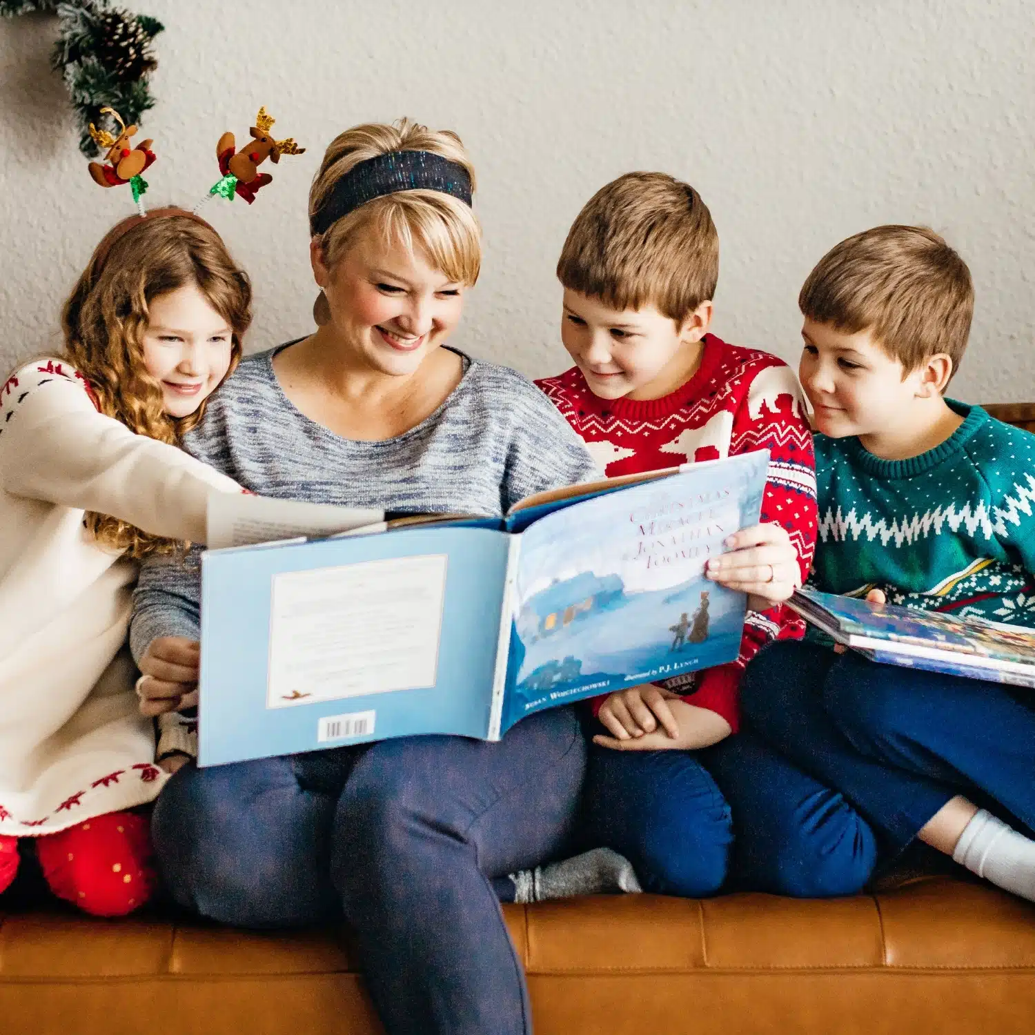 Christmas 7 - Read-Aloud Revival ® with Sarah Mackenzie Family of four on a sofa reading a large picture book together, wearing Christmas sweaters and holiday accessories.