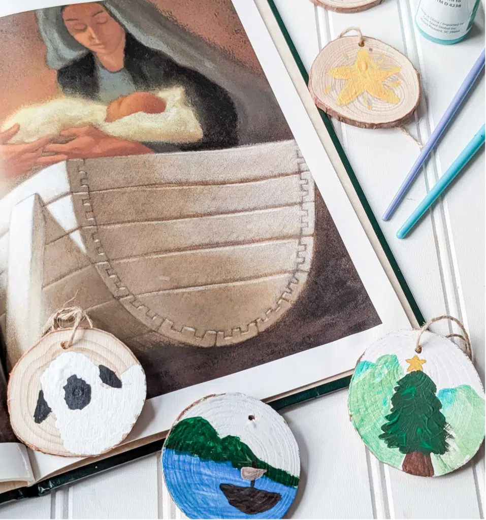 Nativity - Read-Aloud Revival ® with Sarah Mackenzie Assorted handmade wooden Christmas ornaments with painted scenes (panda, trees, lake) and craft supplies on a white surface with string ties in view.