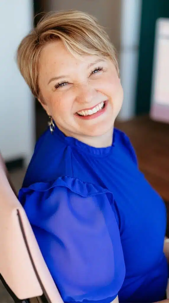 Sarah-Blue-Shirt - Read-Aloud Revival ® with Sarah Mackenzie Smiling woman with short blonde hair wearing a royal blue blouse, seated indoors near a doorway or window.