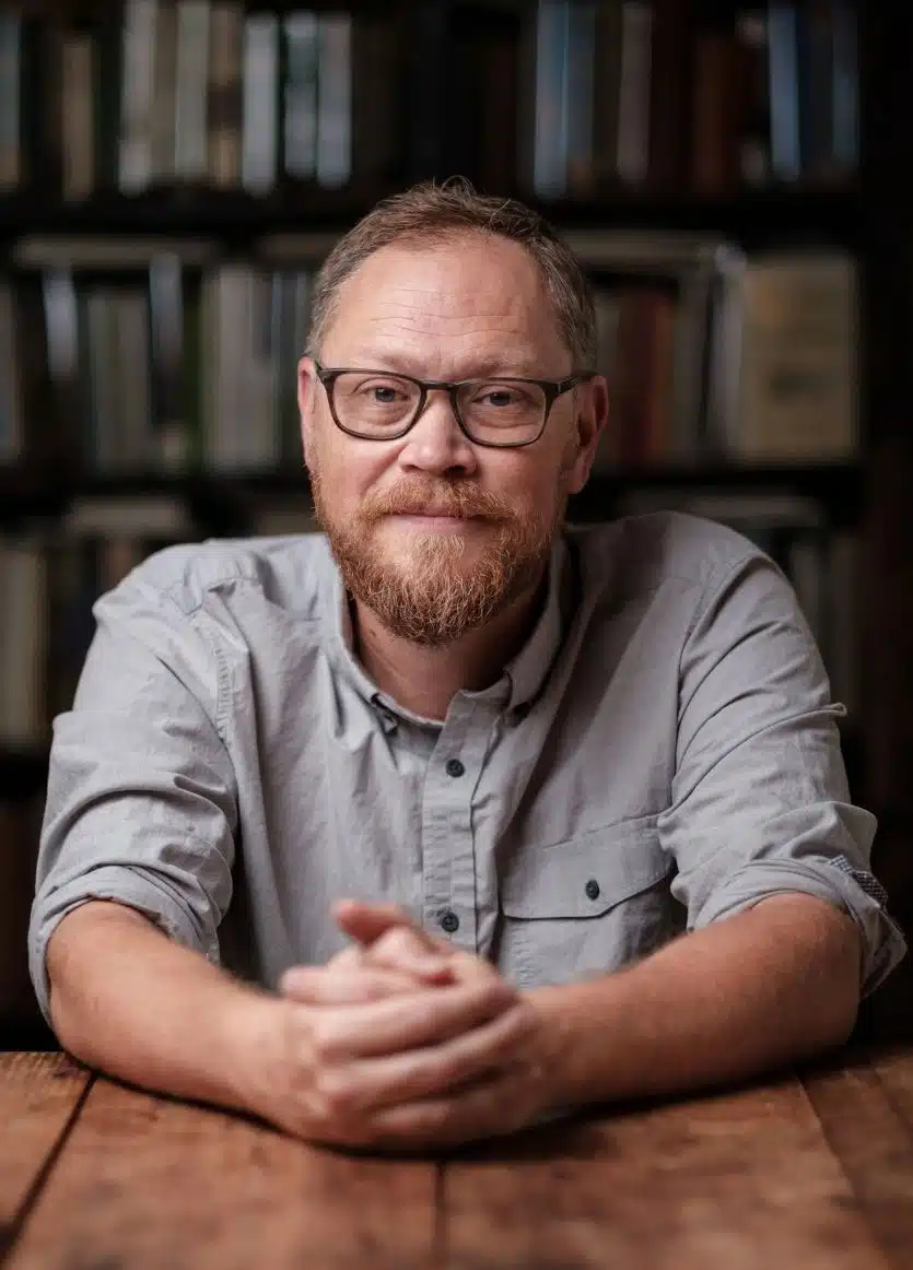 Andrew-Peterson - Read-Aloud Revival ® with Sarah Mackenzie Bearded man with glasses sits at a wooden table, hands folded, in a library backdrop, looking at the camera.