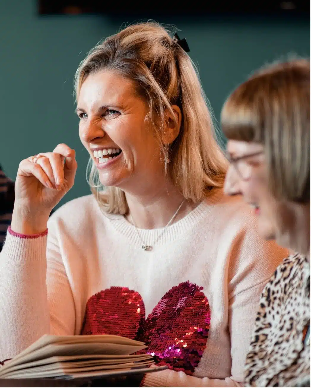 conference - Read-Aloud Revival ® with Sarah Mackenzie Woman laughing while reading a book, wearing a pink sweater with a red sequined heart.