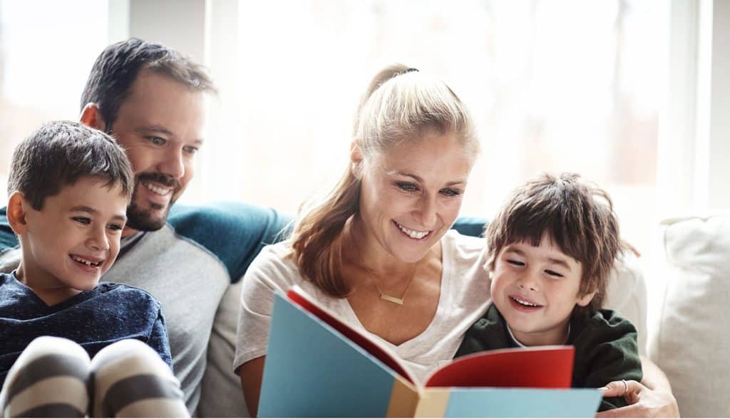 Family of four sits on a couch, reading a colorful book together and smiling.