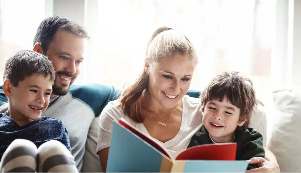 family-book-club-free-guide-2 - Read-Aloud Revival ® with Sarah Mackenzie Family of four sits on a couch, reading a colorful book together and smiling.