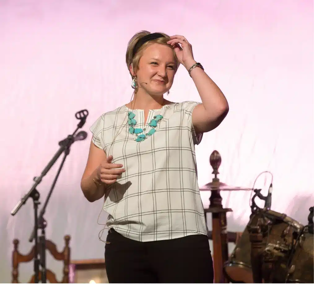 sarah-speaking - Read-Aloud Revival ® with Sarah Mackenzie Woman singer on stage, smiling, wearing a white grid-pattern blouse and turquoise necklace, with a microphone nearby.